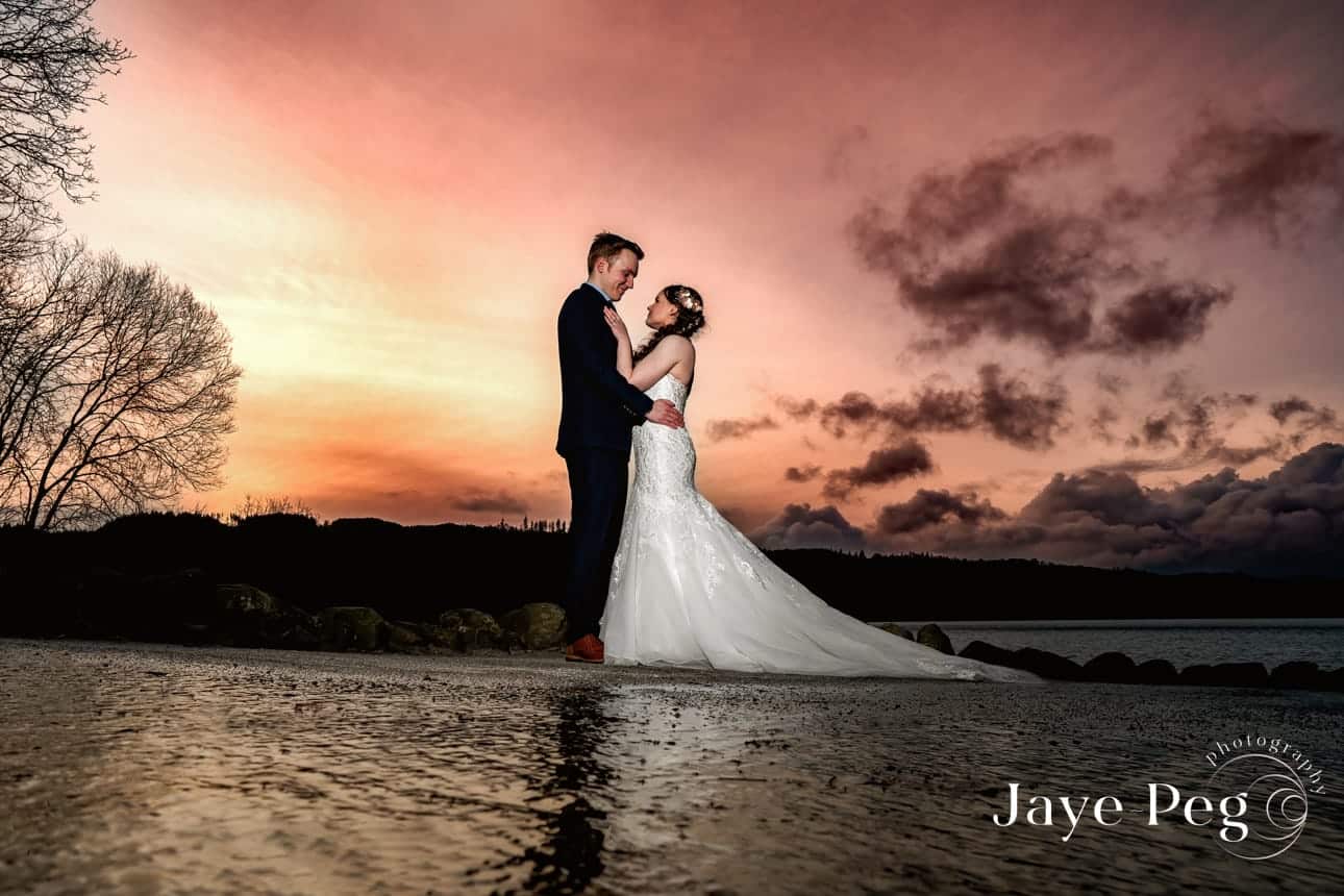 a bride and groom standing on a wet path with a dramatic sunset behind them