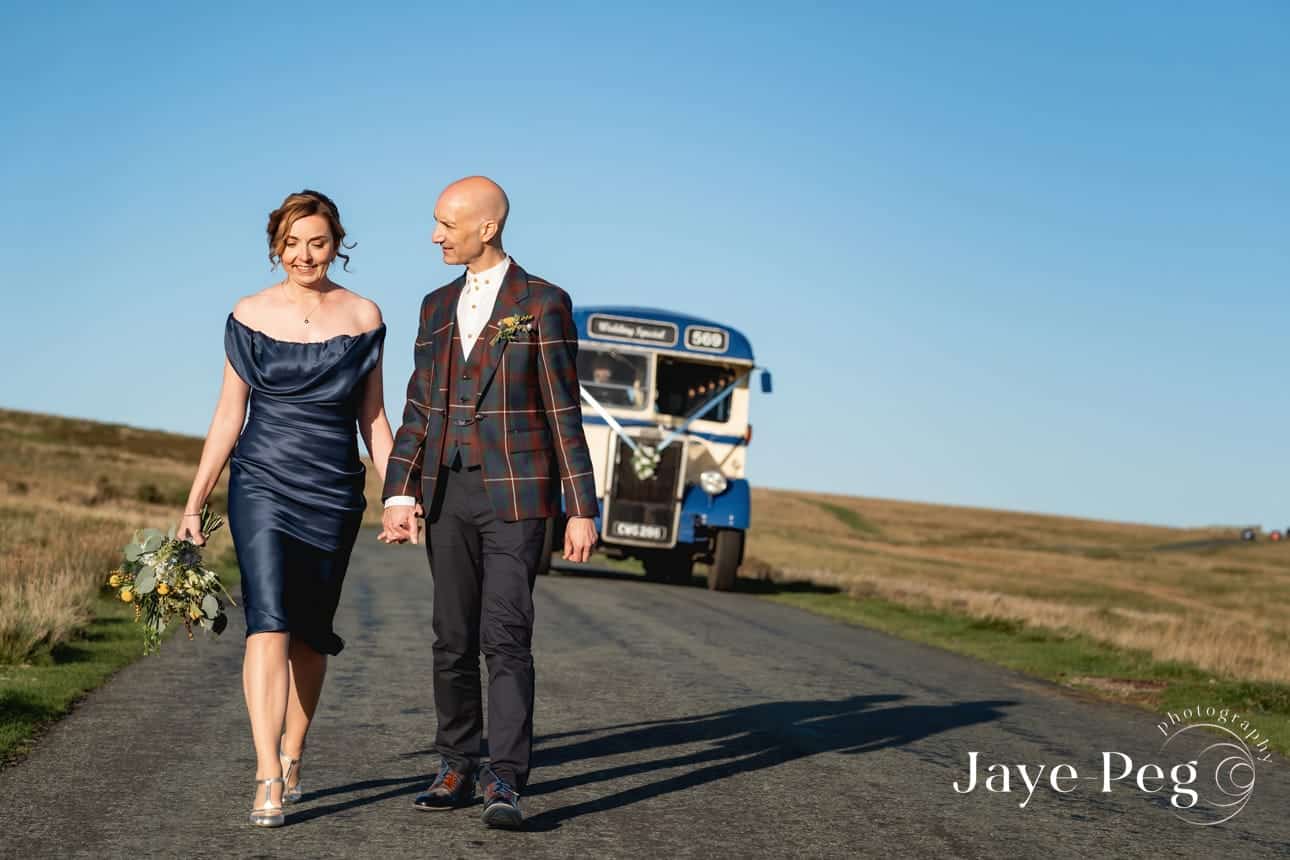 a bride and groom walking along the road at caldbeck fell in cumbria with a vintage bus behind them