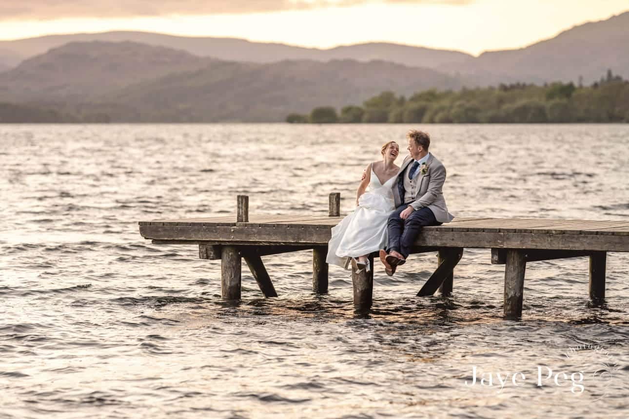 a bride and groom sitting on a jetty during golden hour at windermere