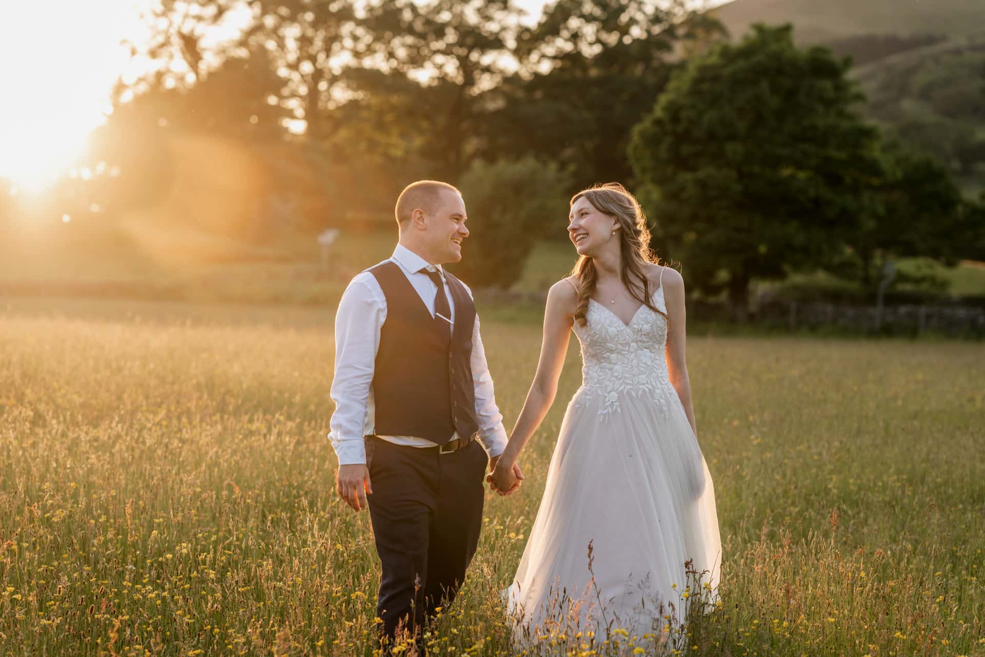 a bride and groom walking through a sunny meadow during golden hour in the lake district