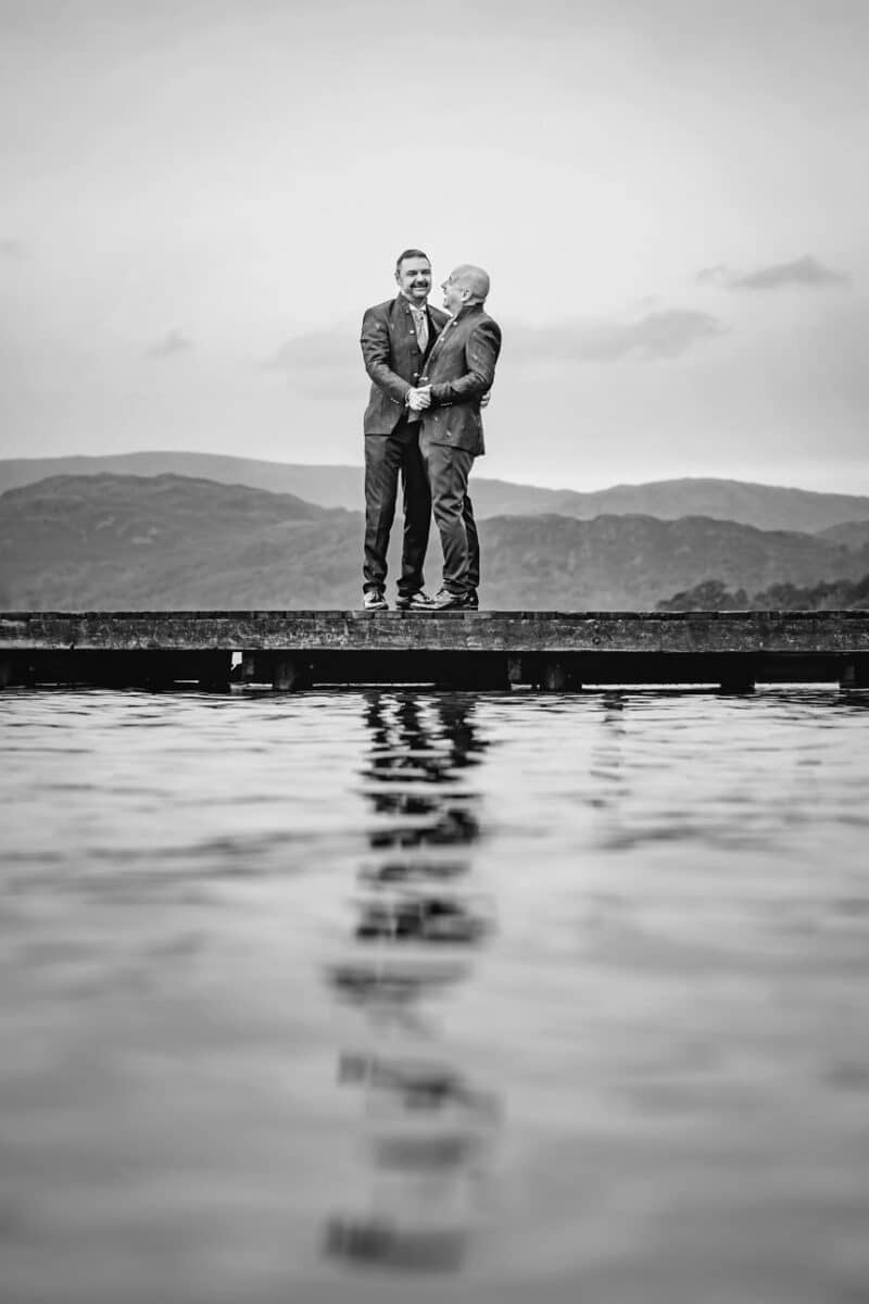 two grooms standing on a jetty over a lake with mountains in the background