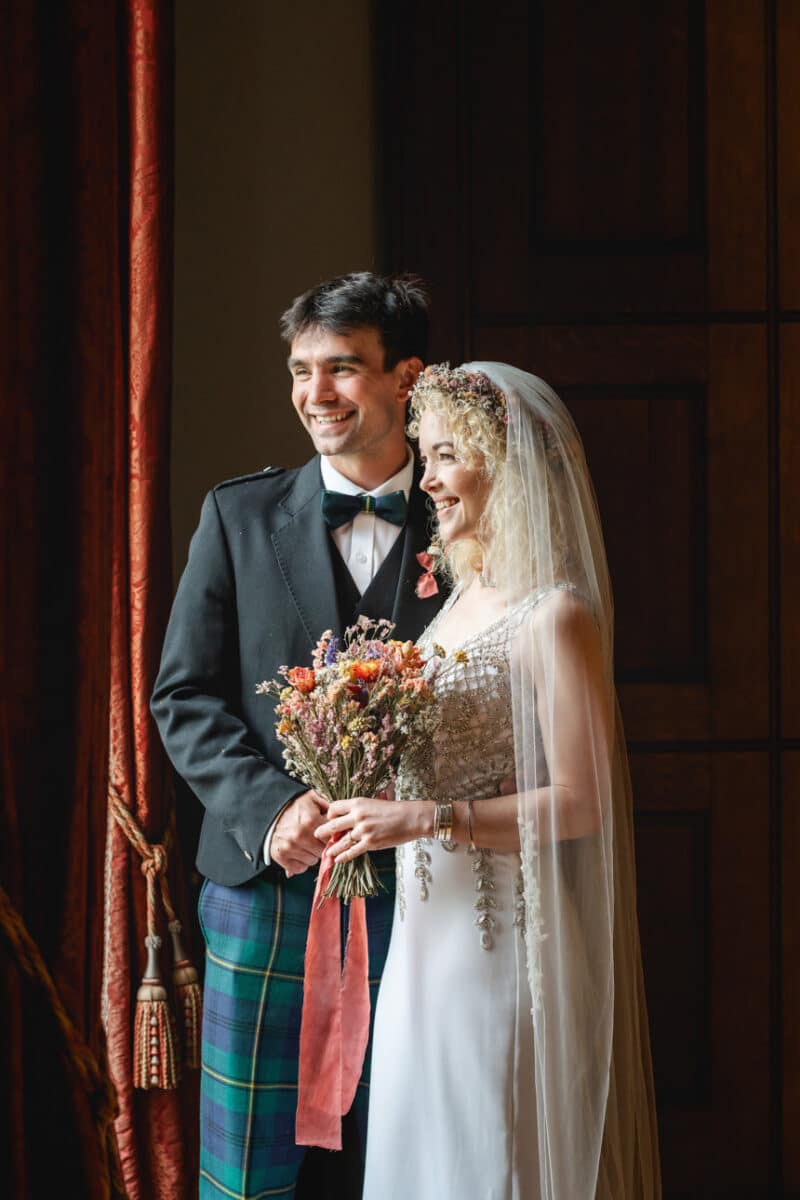 a groom in highland dress looking out of a window with his bride