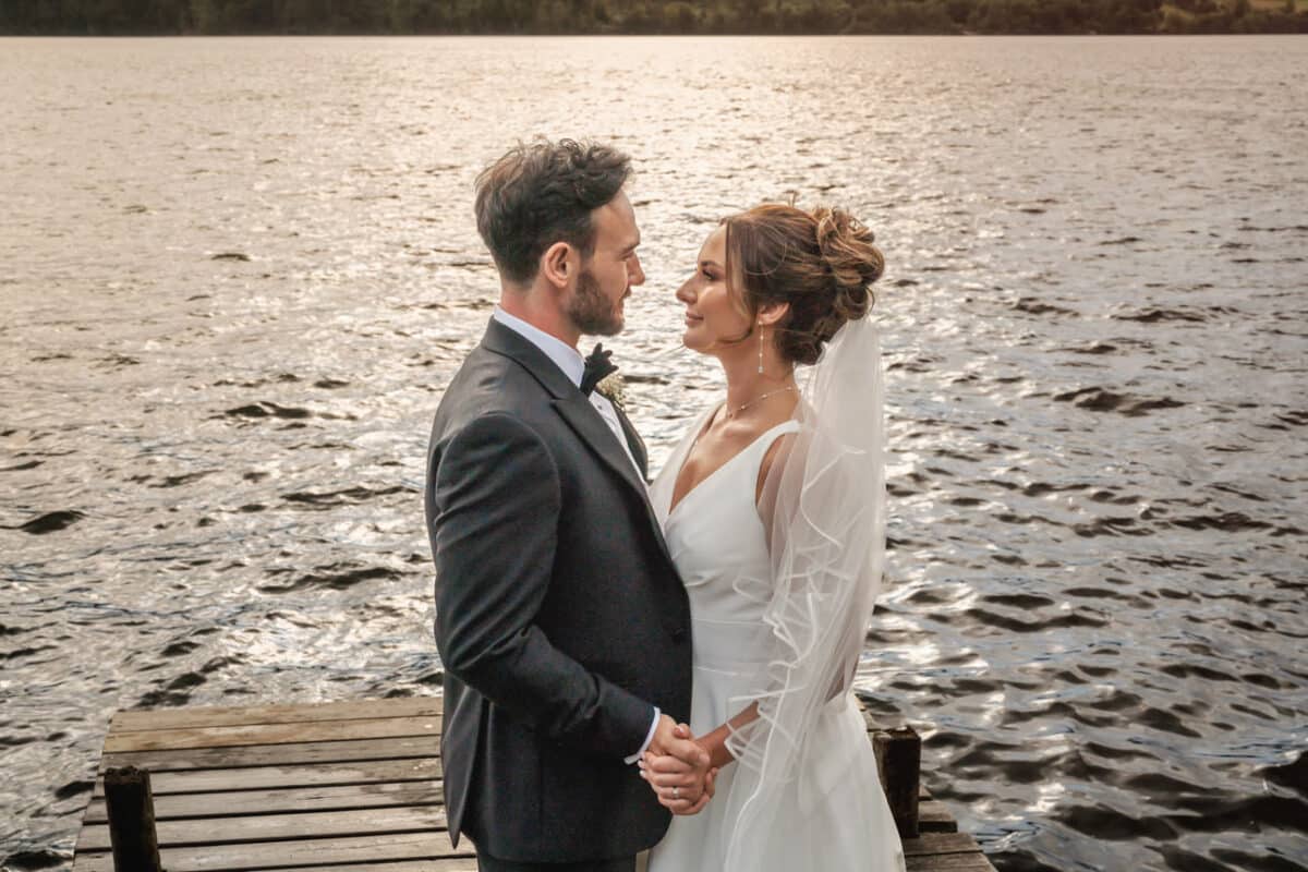 a bride and groom standing on a jetty at sunset with a lake behnind them