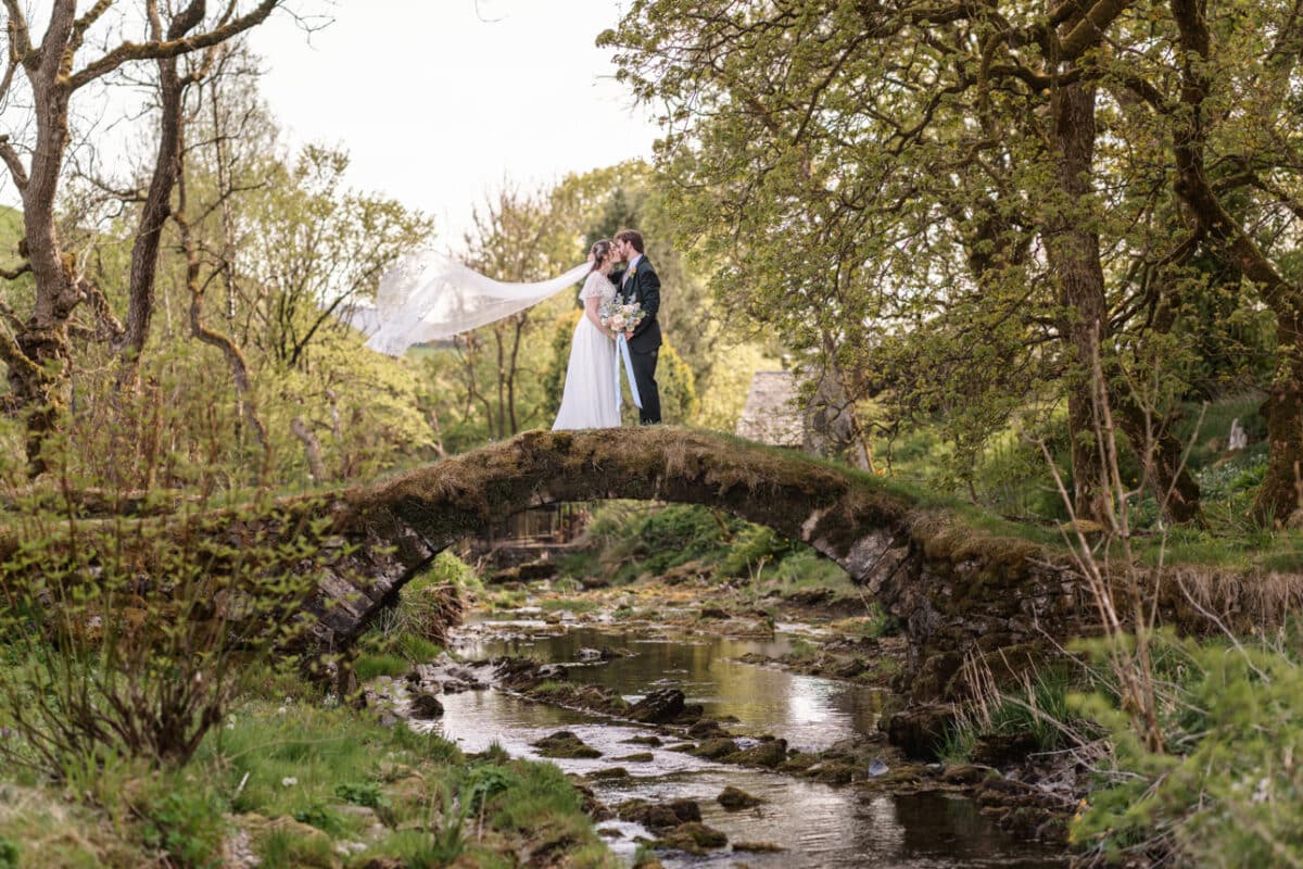a bride and groom on a small stone bridge over a river with the bride's veil blowing behind her