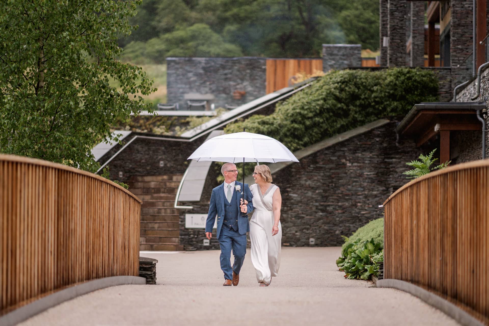 a holding an umbrella and bride and groom walking over a footbridge in the rain