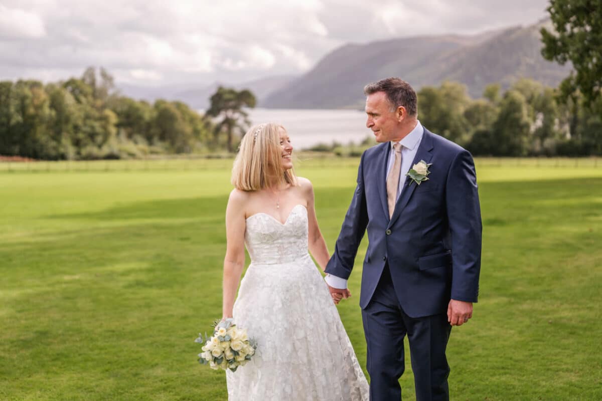 a bride and groom walking on a lawn with bassenthwite lake behind them