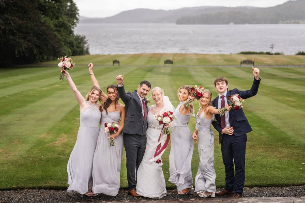 a bride and groom with their wedding party celebrating on a lawn with windermere and the lake district fells in the background