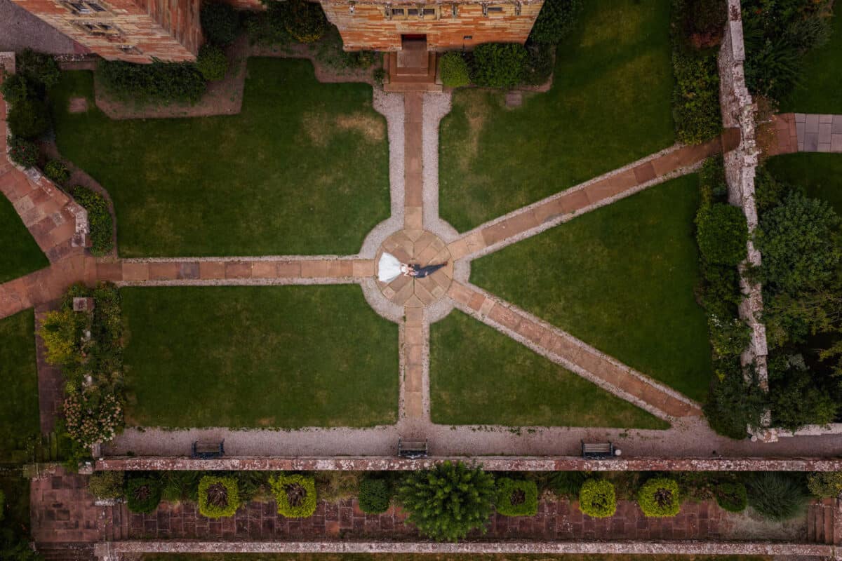 a drone photo of a bride and groom lying on the ground where several footpaths join together in landscaped gardens