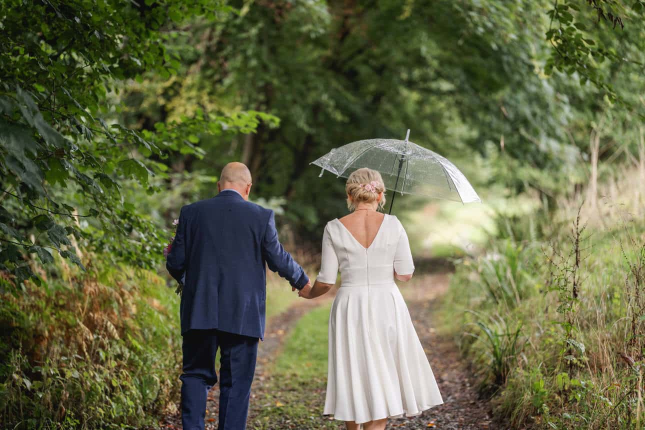 a bride holding an umbrella and a groom walking away down a lane in the countryside