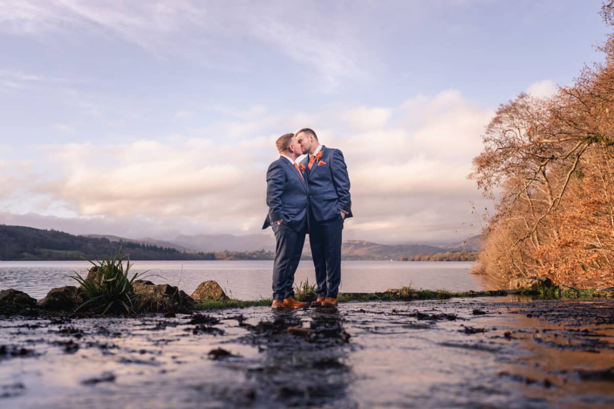 two grooms on a wet path with a lake and the lake district fells in the background